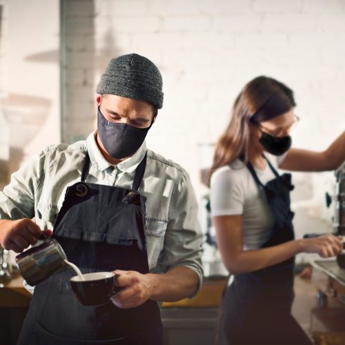 Baristas en una cafetería moderna, uno vertiendo leche en una taza y la otra operando una máquina de espresso, ambos usando mascarillas.